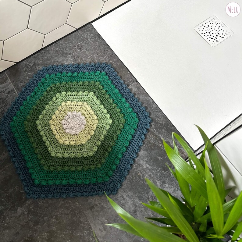 hexagon shaped bath mat made with crocheted cotton yarn using bobble stitch and tr crochet in tones of green. shown in place in a shower room with a plant