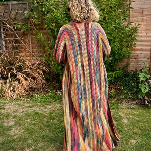 Può includere: Cardigan lungo all'uncinetto colorato con strisce verticali nei toni del rosso, arancione, giallo, verde e blu. Il cardigan è indossato all'aperto in un giardino, con una staccionata di legno e verde sullo sfondo. L'indumento ha maniche lunghe e uno spacco sul fondo.