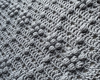 Op de afbeelding: Een close-up van een grijs gehaakt deken met een textuur van daisy-steek. Het deken is gemaakt van zacht garen en heeft een delicate, vrouwelijke uitstraling. De tekst "Crazy Little Daisy Blanket by Melli Crochet" is zichtbaar in de rechterbenedenhoek van de afbeelding.