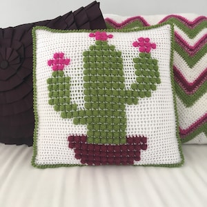 May include: White crocheted pillow with a green cactus design and pink flowers. The cactus is in a burgundy pot.
