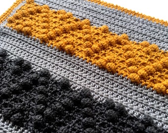 May include: A crocheted blanket with stripes of gray, black, and yellow yarn. The yellow stripe features a textured stitch that creates small bumps.