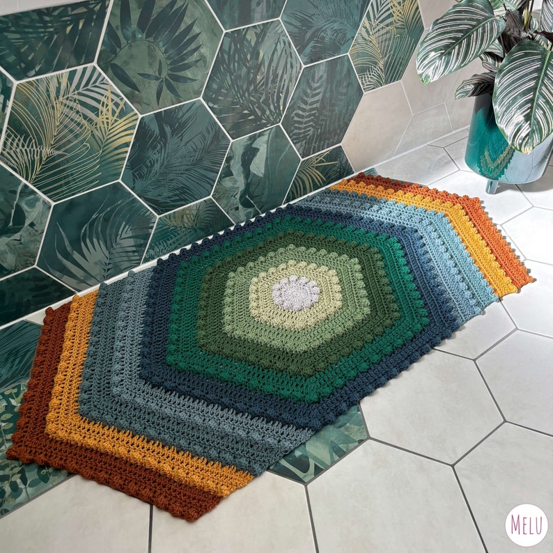 extended hexagon shaped bath mat made with crocheted cotton yarn using bobble stitch and tr crochet in tones of green and orange. shown in place in a bath room with a plant