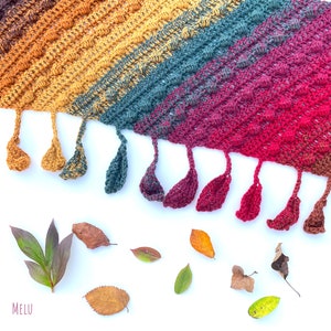 May include: A handmade crochet shawl in a gradient of colors, transitioning from gold to teal, then to burgundy. The shawl has a textured stitch pattern and is adorned with matching tassels. Various leaves are scattered around the shawl on a white background. The word "melu" is in the bottom left corner.