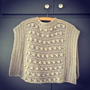 Puede incluir: Un poncho de crochet beige con un patrón texturizado de pequeños círculos. El poncho está colgado en una percha de madera sobre un fondo gris.