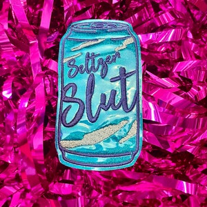 May include: A teal and purple embroidered patch in the shape of a seltzer can. The patch features the words "Seltzer Slut" in a cursive font. The background is a bright pink, metallic, tinsel-like material.