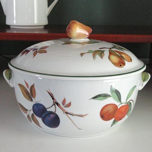 May include: White ceramic serving bowl with lid, featuring a fruit design of plums and pears. The bowl has a green trim and handles. The lid is topped with a pear-shaped knob. A white teapot is visible in the background.