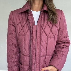 May include: A mauve quilted jacket with a collared neckline and a full-length zipper. The jacket features a diamond pattern and a pocket. A white t-shirt is visible underneath the jacket.
