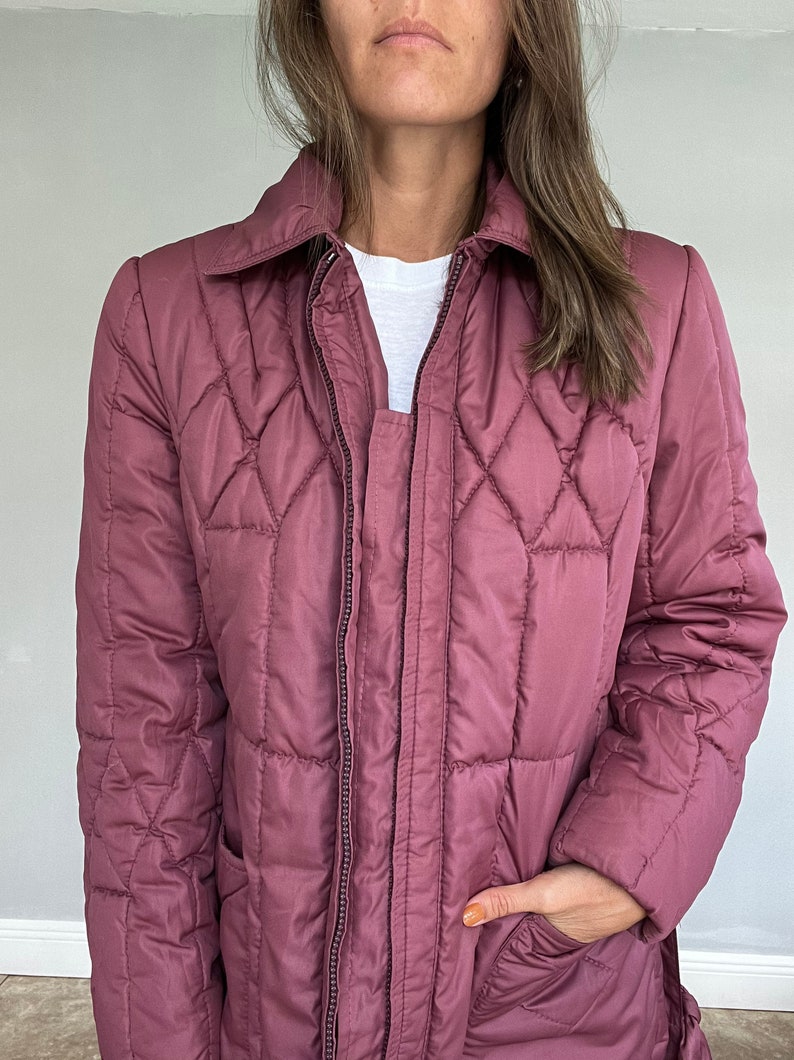 May include: A mauve quilted jacket with a collared neckline and a full-length zipper. The jacket features a diamond pattern and a pocket. A white t-shirt is visible underneath the jacket.