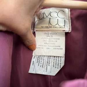 May include: Close-up of a label from a garment, held by a hand. The label is white with black text, including "avec tu" and "THE STERLING STALL GROUP." The garment's lining is a deep burgundy color.