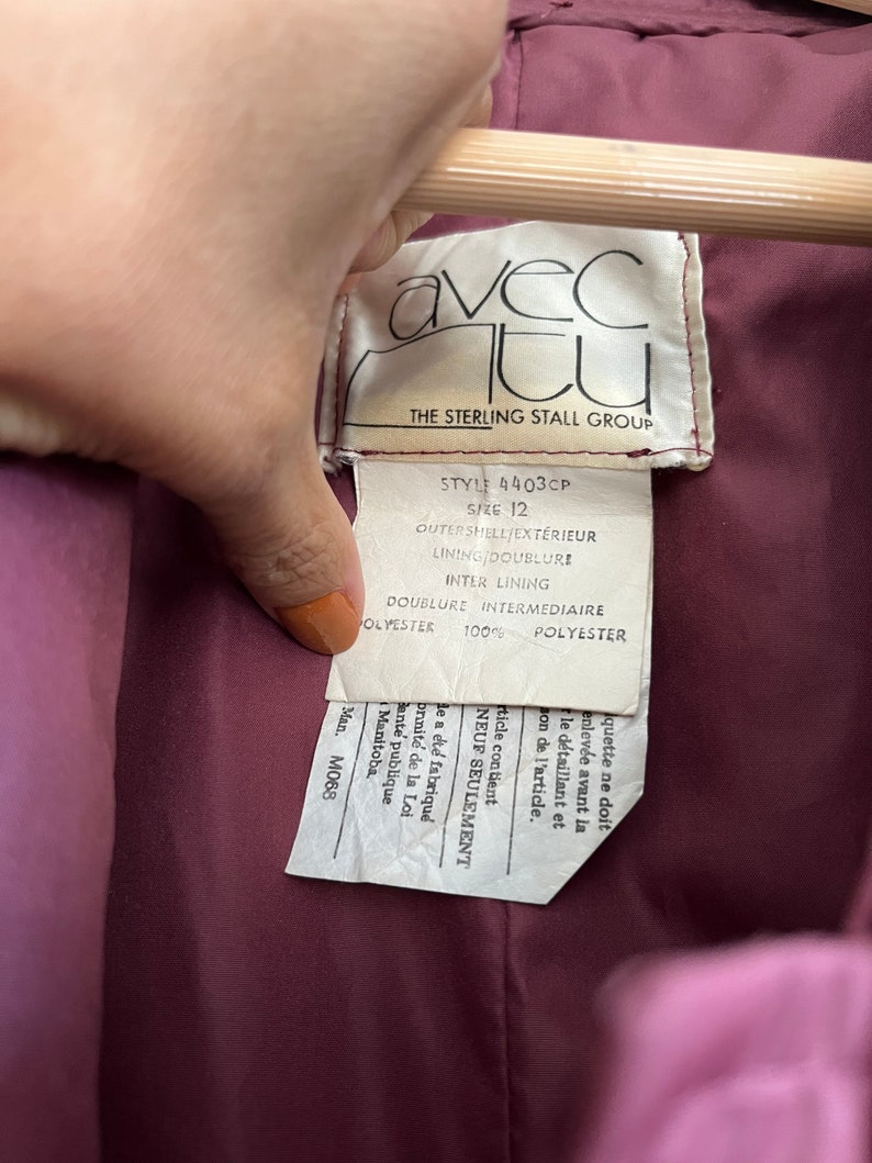 May include: Close-up of a label from a garment, held by a hand. The label is white with black text, including "avec tu" and "THE STERLING STALL GROUP." The garment's lining is a deep burgundy color.