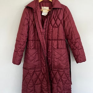 May include: A burgundy quilted coat with a diamond pattern, long sleeves, and a belt. The coat has a collar, two front pockets, and a zipper closure. The coat is hanging on a wooden hanger.
