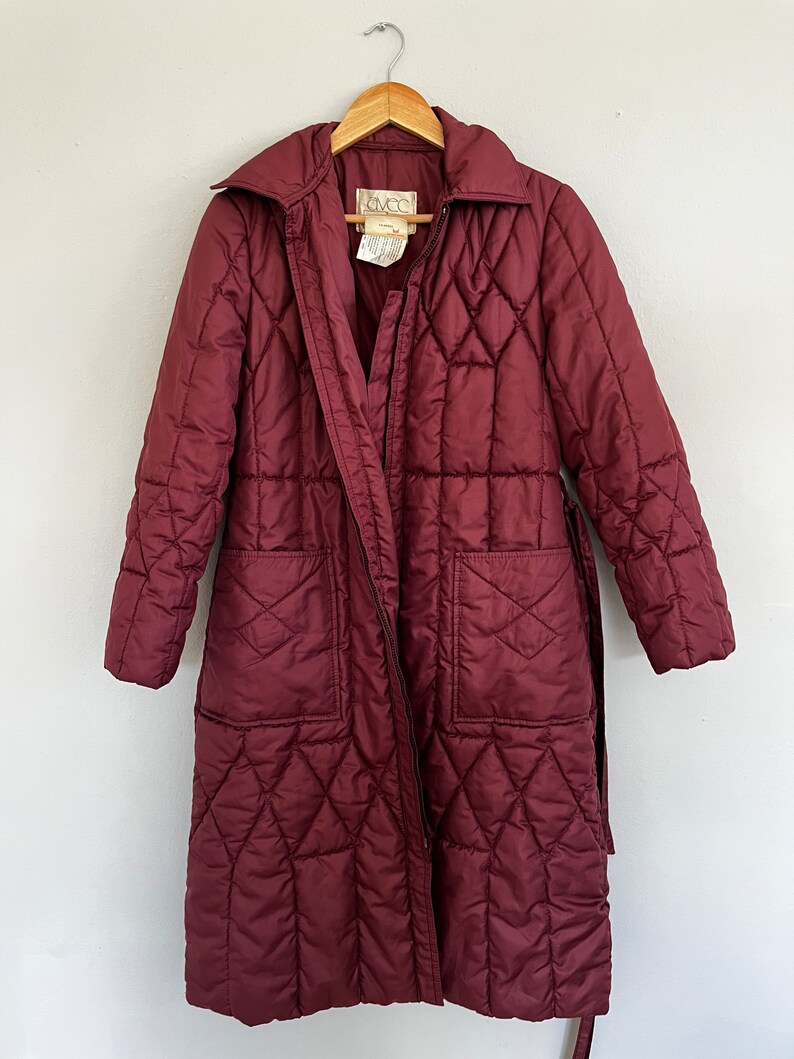 May include: A burgundy quilted coat with a diamond pattern, long sleeves, and a belt. The coat has a collar, two front pockets, and a zipper closure. The coat is hanging on a wooden hanger.