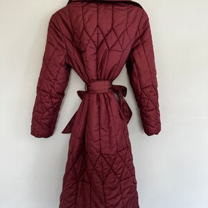 May include: A burgundy quilted coat with a diamond pattern, hanging on a wooden hanger. The coat features a wide collar, long sleeves, and a matching belt tied at the waist. The coat is knee-length.