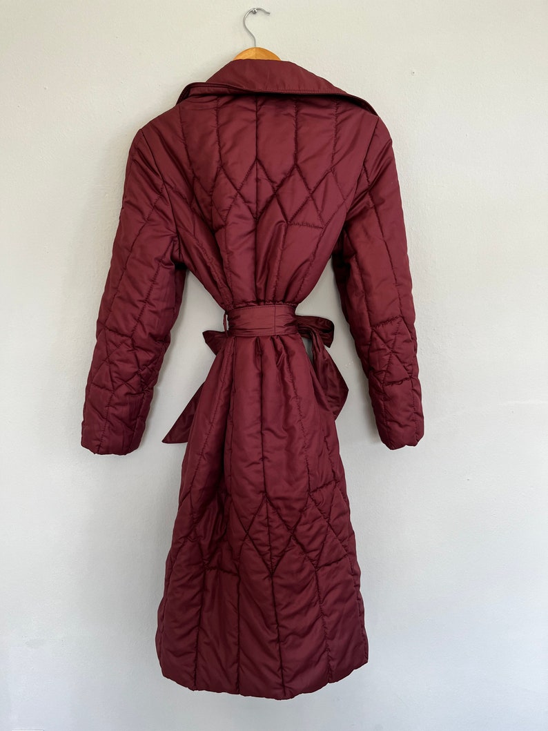 May include: A burgundy quilted coat with a diamond pattern, hanging on a wooden hanger. The coat features a wide collar, long sleeves, and a matching belt tied at the waist. The coat is knee-length.