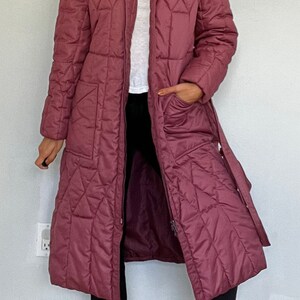 May include: A long, quilted coat in a muted rose color. The coat features a diamond pattern, a collar, and a belt. It has a zipper closure and pockets. The person is wearing black pants and black ankle boots.
