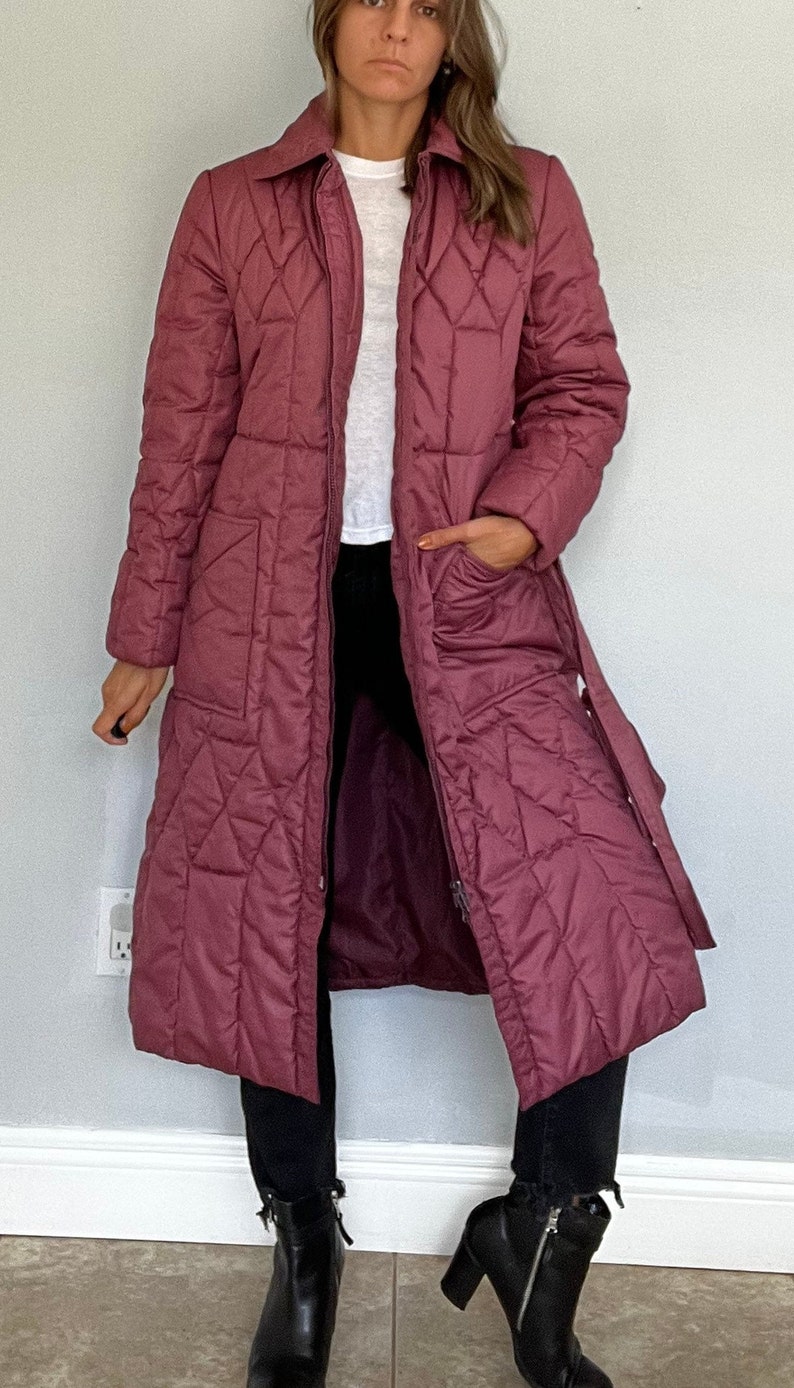 May include: A long, quilted coat in a muted rose color. The coat features a diamond pattern, a collar, and a belt. It has a zipper closure and pockets. The person is wearing black pants and black ankle boots.
