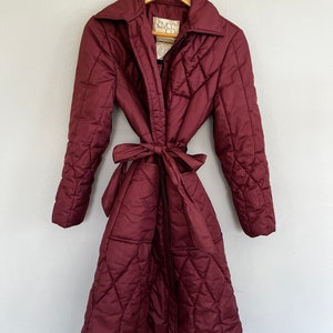 May include: A burgundy quilted coat with a tie belt, hanging on a wooden hanger. The coat has a wide collar and a diamond pattern. The coat is a long, full-length style.