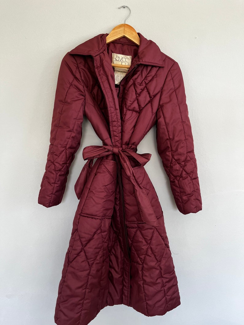 May include: A burgundy quilted coat with a tie belt, hanging on a wooden hanger. The coat has a wide collar and a diamond pattern. The coat is a long, full-length style.