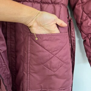 May include: Close-up of a quilted, mauve-colored coat with a hand in the pocket. The coat features a diamond pattern and a visible pocket. A gold bracelet is on the wrist.