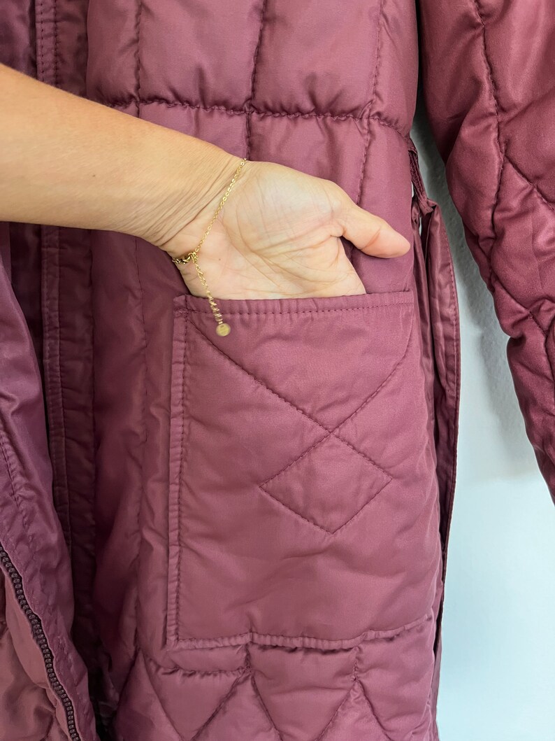 May include: Close-up of a quilted, mauve-colored coat with a hand in the pocket. The coat features a diamond pattern and a visible pocket. A gold bracelet is on the wrist.