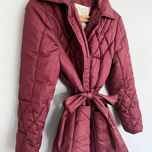 May include: A burgundy quilted coat with a matching tie belt. The coat has a collared neckline and a zipper closure. The coat is hanging on a wooden hanger. The label reads "AVEC".