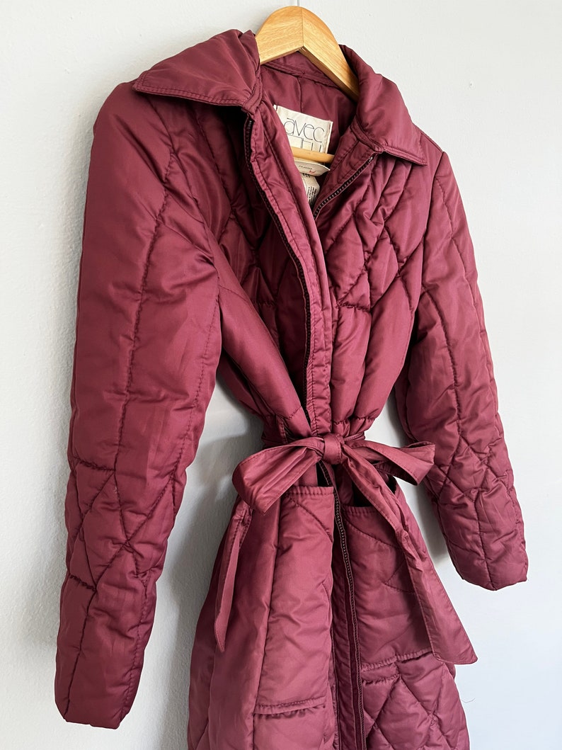 May include: A burgundy quilted coat with a matching tie belt. The coat has a collared neckline and a zipper closure. The coat is hanging on a wooden hanger. The label reads "AVEC".