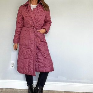 May include: A long, quilted coat in a dusty rose color. The coat features a wrap-around design with a matching belt, a collar, and pockets. The coat has a diamond pattern and is worn with black pants and boots.