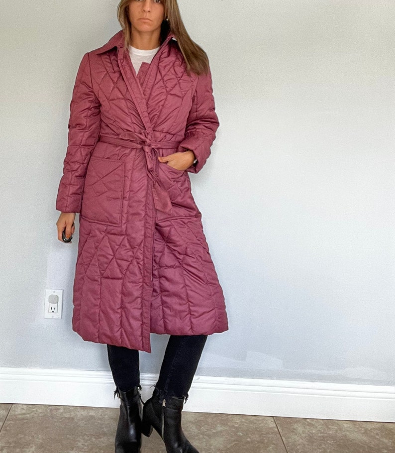 May include: A long, quilted coat in a dusty rose color. The coat features a wrap-around design with a matching belt, a collar, and pockets. The coat has a diamond pattern and is worn with black pants and boots.