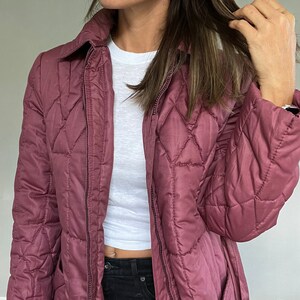 May include: A quilted, mauve-colored jacket with a collared neckline and a diamond pattern. The jacket has a zipper closure and pockets. A white t-shirt is visible underneath. The model is wearing black jeans.