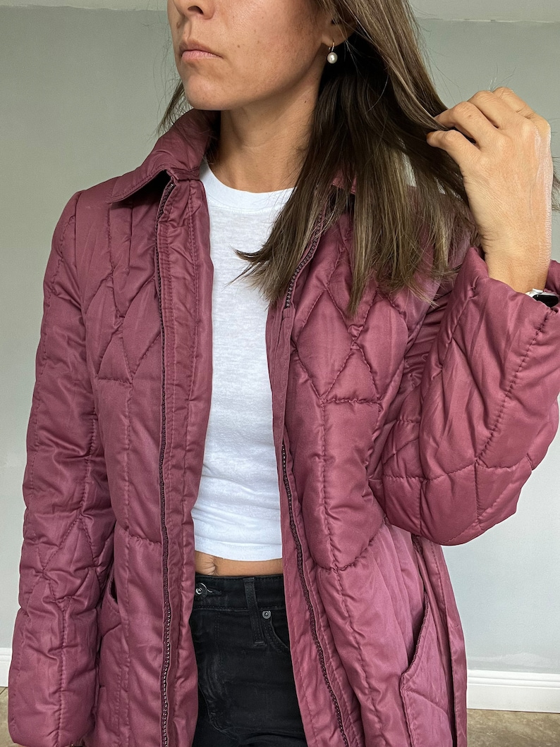 May include: A quilted, mauve-colored jacket with a collared neckline and a diamond pattern. The jacket has a zipper closure and pockets. A white t-shirt is visible underneath. The model is wearing black jeans.