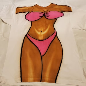 Pode incluir: Camiseta branca com uma imagem impressa de uma pessoa de biquíni rosa. A pessoa tem pele bronzeada e é representada do peito para baixo.