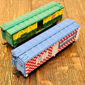 May include: Two toy train cars. One is green with yellow accents and the text "CNW 15040". The other is blue with red and white checkered patterns and the text "Ralston Purina Co".