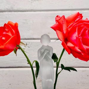 May include: A frosted glass figurine of a woman stands between two vibrant red roses. The roses are in full bloom, with green stems and leaves. The background is a white, horizontal wood-paneled wall.