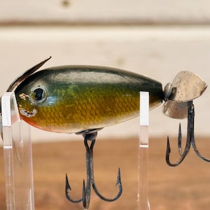 May include: A vintage fishing lure with a green and gold body, silver propeller, and three black treble hooks.