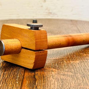 May include: A wooden marking gauge tool featuring a rectangular head and a long cylindrical beam. The tool includes a metal thumbscrew for adjustment on top and a circular metal end cap with a central screw on the side of the wooden beam.