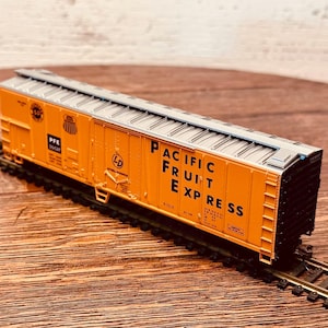 May include: A model train car in orange with the text "PACIFIC FRUIT EXPRESS" on the side. The car is on a model train track. The car has a silver roof and black ends.