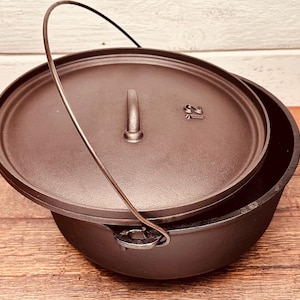 May include: A black cast iron Dutch oven with a lid and a metal handle. The lid has a handle and the number "12" is stamped on the lid. The pot has a metal handle for carrying. The pot is sitting on a wooden surface.
