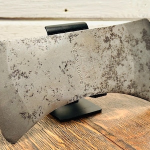 May include: A double-bit axe head made of weathered steel with visible rust spots. The metal surface features the stamped text VULCAN KELLY WORKS. The axe head is held in place by a black metal stand, showcasing two sharp, curved cutting edges.