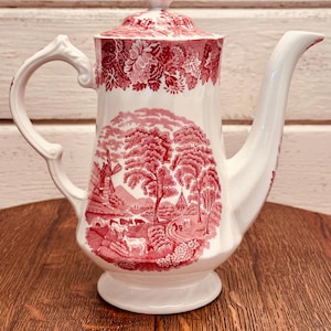 May include: A white ceramic teapot with a red pastoral scene design. The teapot features a curved handle, a long spout, and a lid with a decorative finial. The design includes a windmill, trees, and figures.