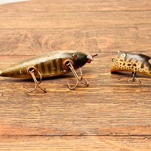 May include: Two fishing lures, one with a brown and green striped body and the other with a brown and black spotted body. Both lures have multiple hooks and are made of wood.