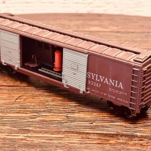 May include: A maroon model train boxcar with the name "SYLVANIA" and the number 83247 printed on the side. The boxcar has a partially open door, revealing interior details and a red cylinder. The boxcar is on a wooden surface.