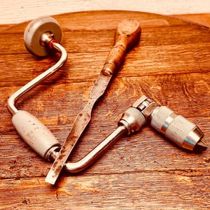 May include: An assortment of vintage woodworking tools, including a brace and bit, a chisel, and a hand drill. The tools are made of metal and wood, with signs of wear and use. The brace and bit has a gray handle and a curved metal arm. The chisel has a wooden handle and a metal blade. The hand drill has a textured metal handle.