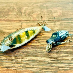 May include: Two vintage fishing lures. One is a white and yellow lure with green stripes and a silver propeller. The other is a black and white lure with a silver propeller. Both have hooks.