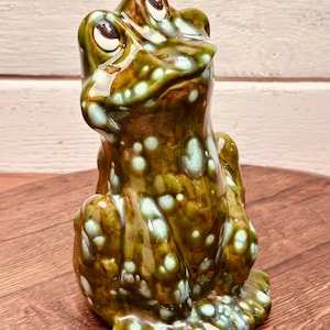 May include: A ceramic frog figurine with a glossy finish. The frog is primarily green and brown with white and light blue spots. The frog is sitting upright with its eyes looking up.