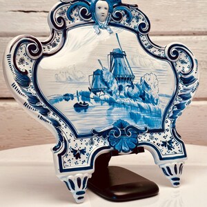 May include: A decorative ceramic plaque featuring a blue and white windmill scene. The plaque has an ornate, scrolled border and is supported by a black stand. The artwork depicts windmills, water, and a small boat.