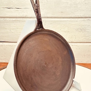 May include: A round, cast iron griddle with a long handle and a loop for hanging. The griddle is a dark brown color and has a slightly textured surface. The handle is attached to the griddle with a seamless transition. The griddle is designed for cooking.