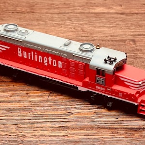 May include: A red and gray model train engine with the word "Burlington" in white lettering on the side. The model has the number 5628 on the side and is set against a wooden background.