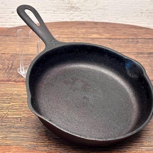 May include: A small, black cast iron skillet with a handle featuring a hole for hanging. The pan has a slightly curved shape and a textured cooking surface. The skillet is designed for stovetop or oven use.