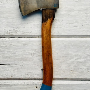 May include: A hand axe with a metallic steel head and a curved wooden handle. The handle features a dark stained upper section, a natural wood grain middle, and a solid blue painted base. Two small silver metal pins are visible on the handle.