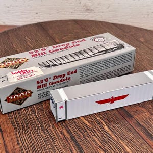 May include: A model train set box and car. The box is gray with the text "52'6" Drop End Mill Gondola" and "Proto 2000 Series." The car is silver with a red bird logo and the number 48.